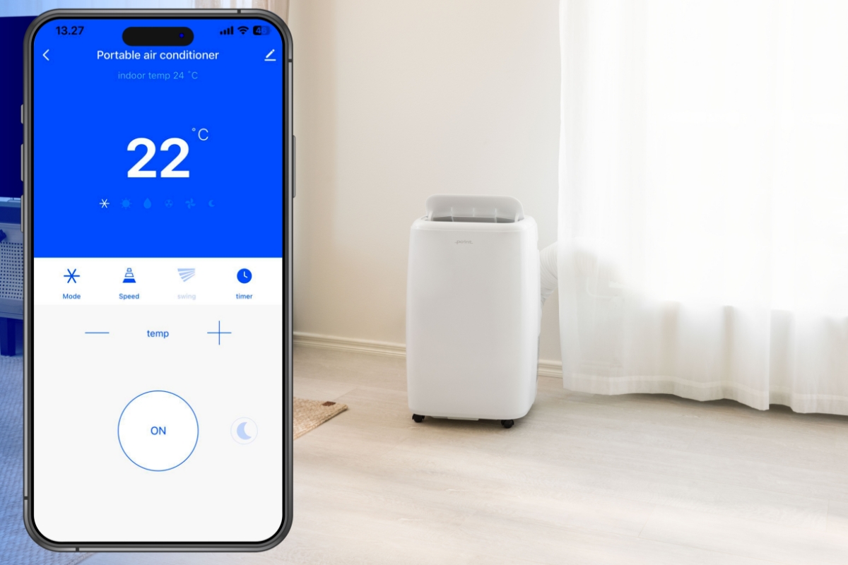 A white Point air conditioner on a light-brown floor in a living room with white curtains and a smartphone on the front showing the SmartLife-app that can be used with the air conditioner