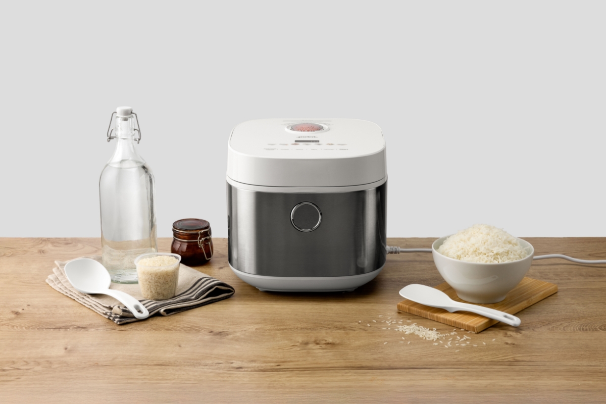 Wide angle image of POINT PORC5010 RICE/MULTI COOKER 5L on wooden countertop, with a cooked bowl of rice next to it. Rice grains are scattered on the counter top next to a jug of water.
