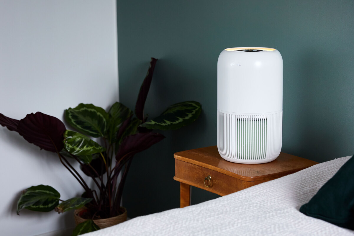 Air purifier on the side table next to a bed and plant from another angle