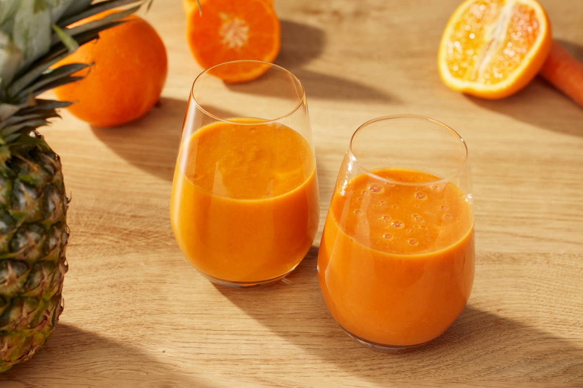 Two small glasses on a wooden table full of orange juice made with Point slowjuicer and a bunch of orange-colored fruits surrounding the glasses