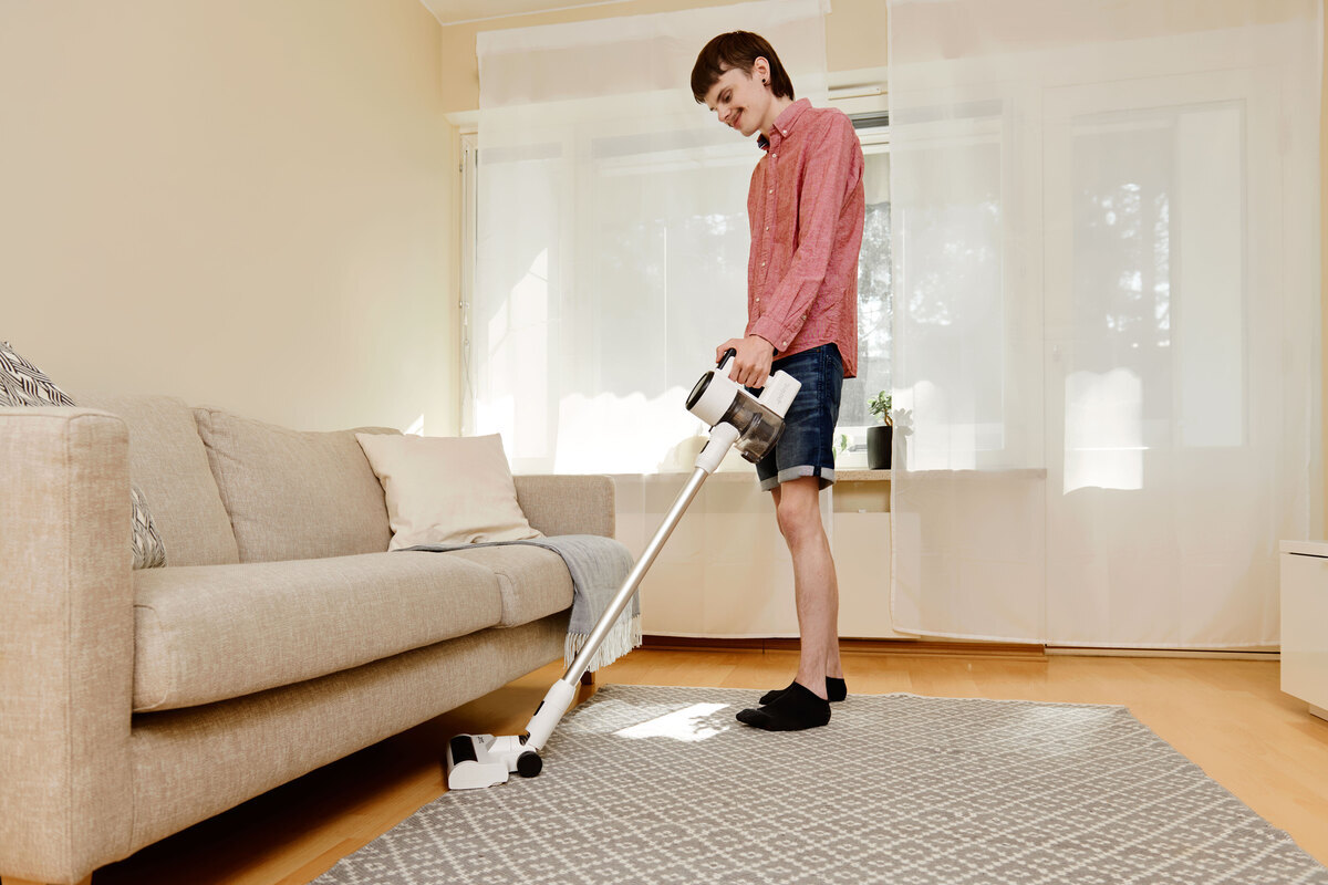 A wide angle image of a person in shorts and a light red shirt vacuuming a grey carpet with the Point Pencil stick vacuum cleaner