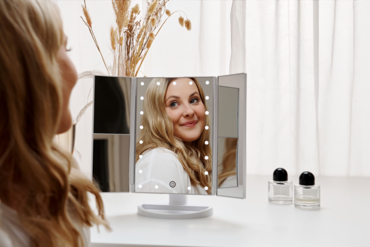 A woman with blonde hair looking at herself in a Nooa Glare make-up mirror with 21 led-bulbs and lots of white light and furniture behind the mirror
