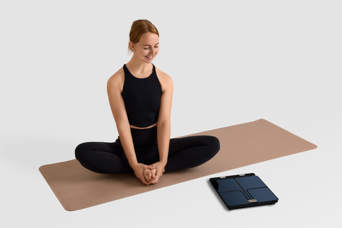 Person in black active wear sitting on a beige yoga mat with the NOOA PHYSIQ SMART PERSONAL SCALE in front of the mat