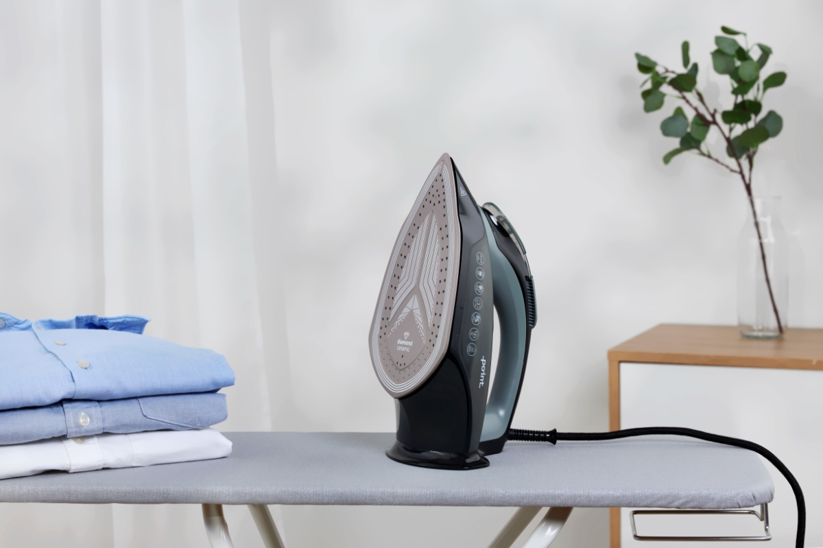 A black Point steam iron in an upright position on a grey ironing board with a stack of blue and white collars next to it and a green plant behind