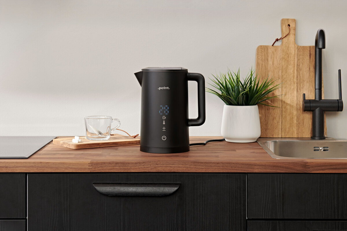 Wide angle image of the black POINT PRO POKED6015E KETTLE on a dark wooden kitchen counter with a plant and a wooden cutting board next to it