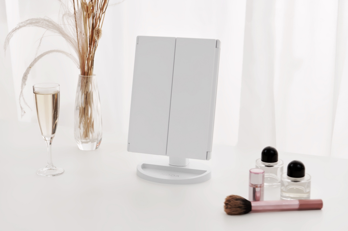 A Nooa Glare make-up mirror standing on a white table with perfumes and a glass of champagne next to it and both its folding mirror closed