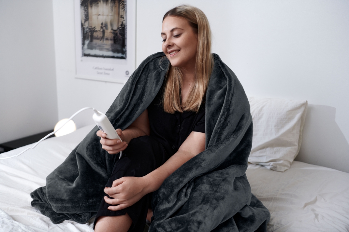 A person sitting on a white bedding with POINT SOOTHING HEAT HEATING BLANKET wrapped around them