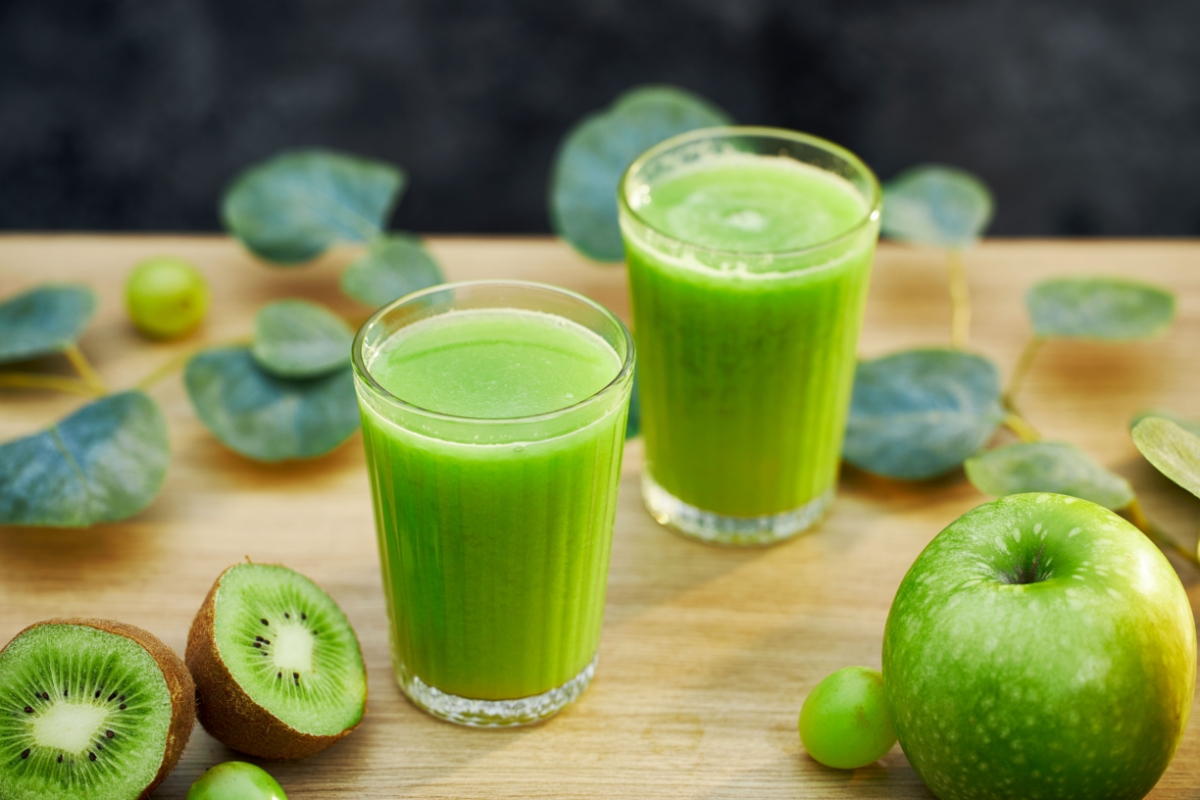 An image of green vegetables and a green smoothie in two see-through glasses on a wooden table