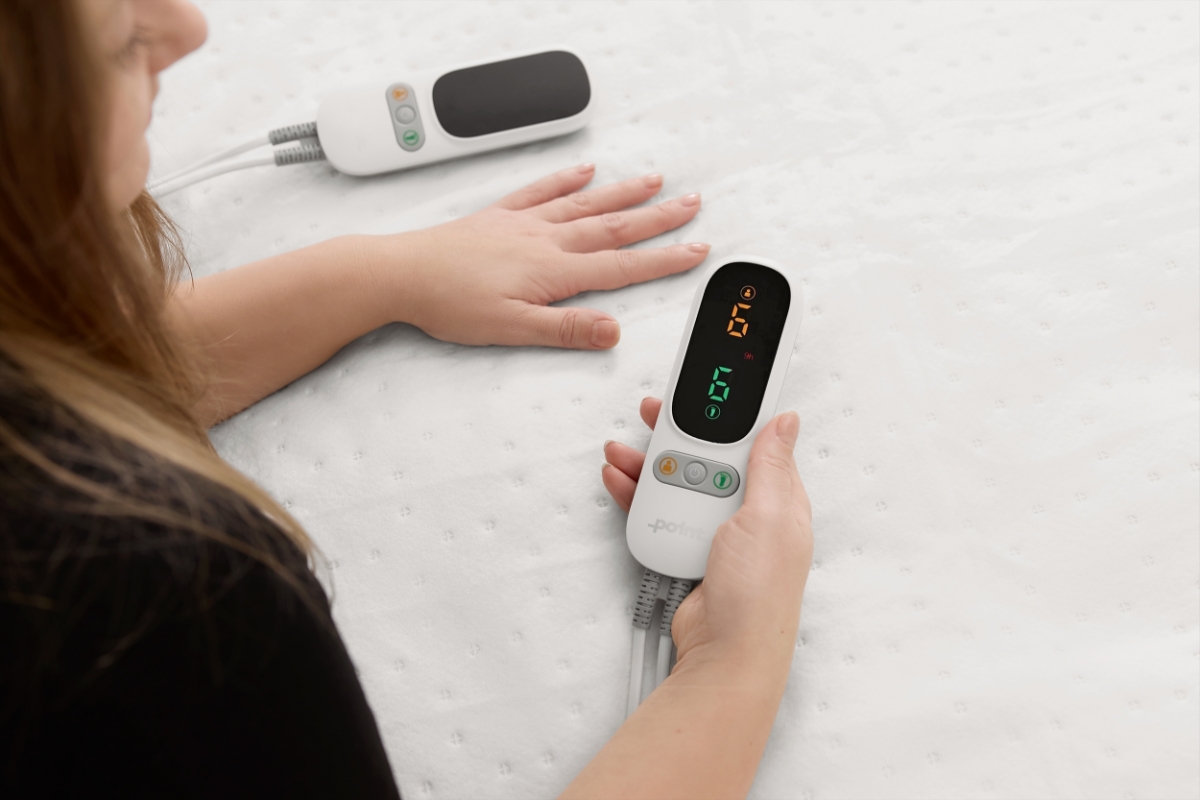 A white Point heated underblanket on the bed and a woman with neat nails and a black shirt holding the controller of the blanket in her hand and the other hand touching the blanket