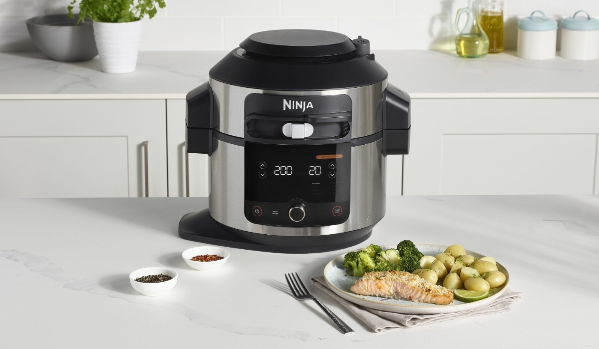A picture of a black and silver Ninja multi cooker standing on a white kitchen table next to a plate full of chicken and green vegetables
