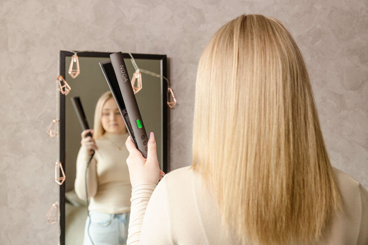 A woman holding a NOOA straightening iron in her hand in front of a mirror