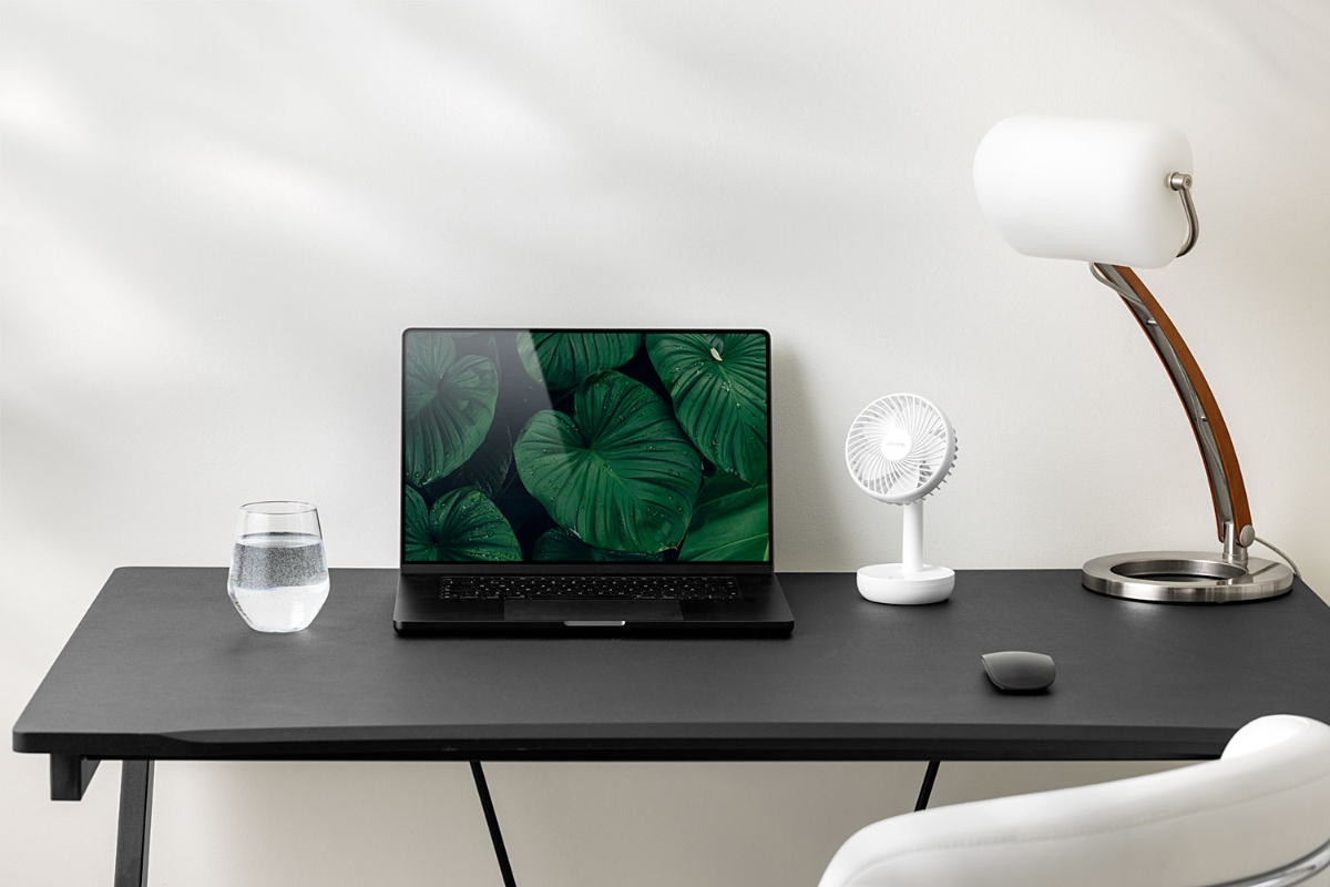 A white and small Point portable fan on a black work desktop next to a black laptop and a white lamp with a white wall behind them