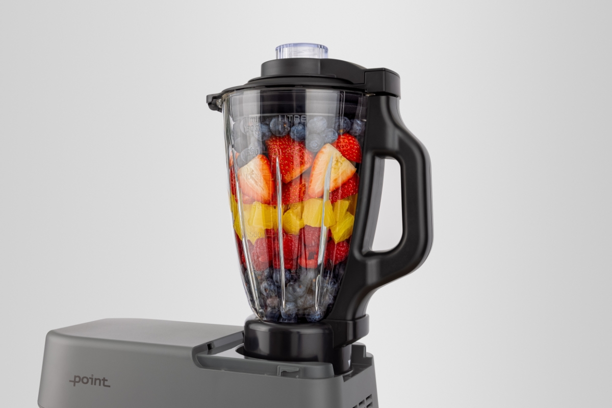Grey-colored Point POKM5010 kitchen machine on a wooden table pictured a bit from the side with the top part only showing and the blender attachment attached to the top filled with fresh berries in red and orange colors