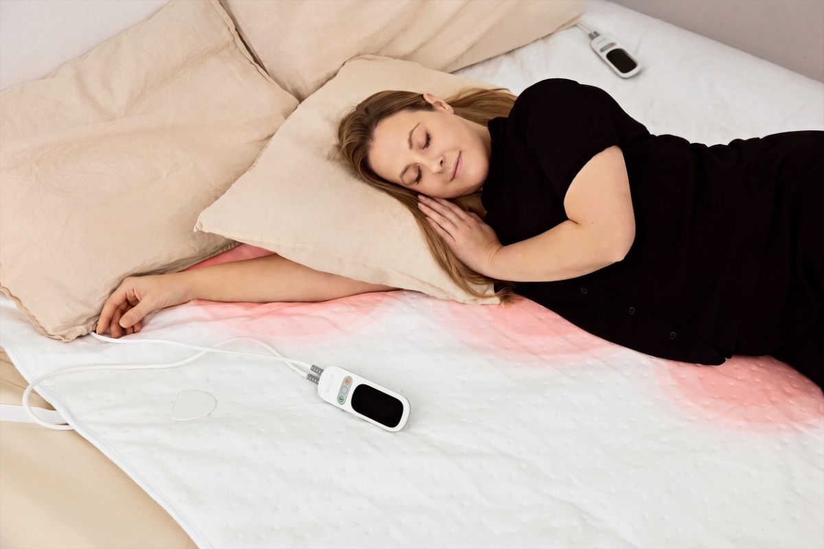 A white Point heated underblanket on a bed with a woman in a black shirt lying on the bed with red dots under her showing the heated areas on the blanket