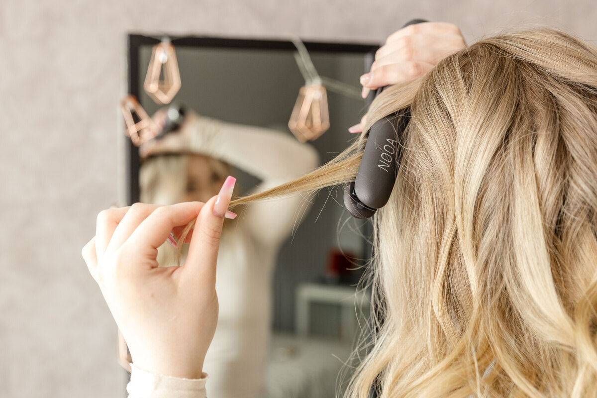 A woman with blonde hair straightening her hair with a dark grey NOOA straightener in front of a mirror