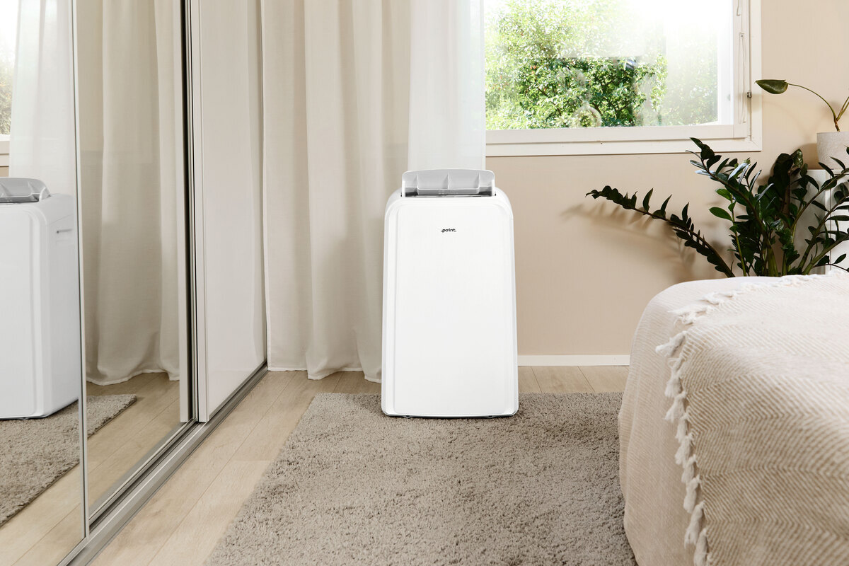 Wide angle image of the POINT PRO POAC8014 air conditioner in front of a beige wall with a green plant and a bed in the right corner