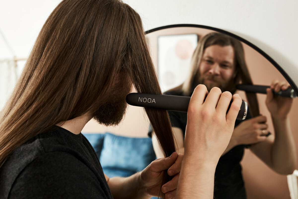 A smiling man styling his long hair with the dark grey NOOA straightener in front of a mirror 