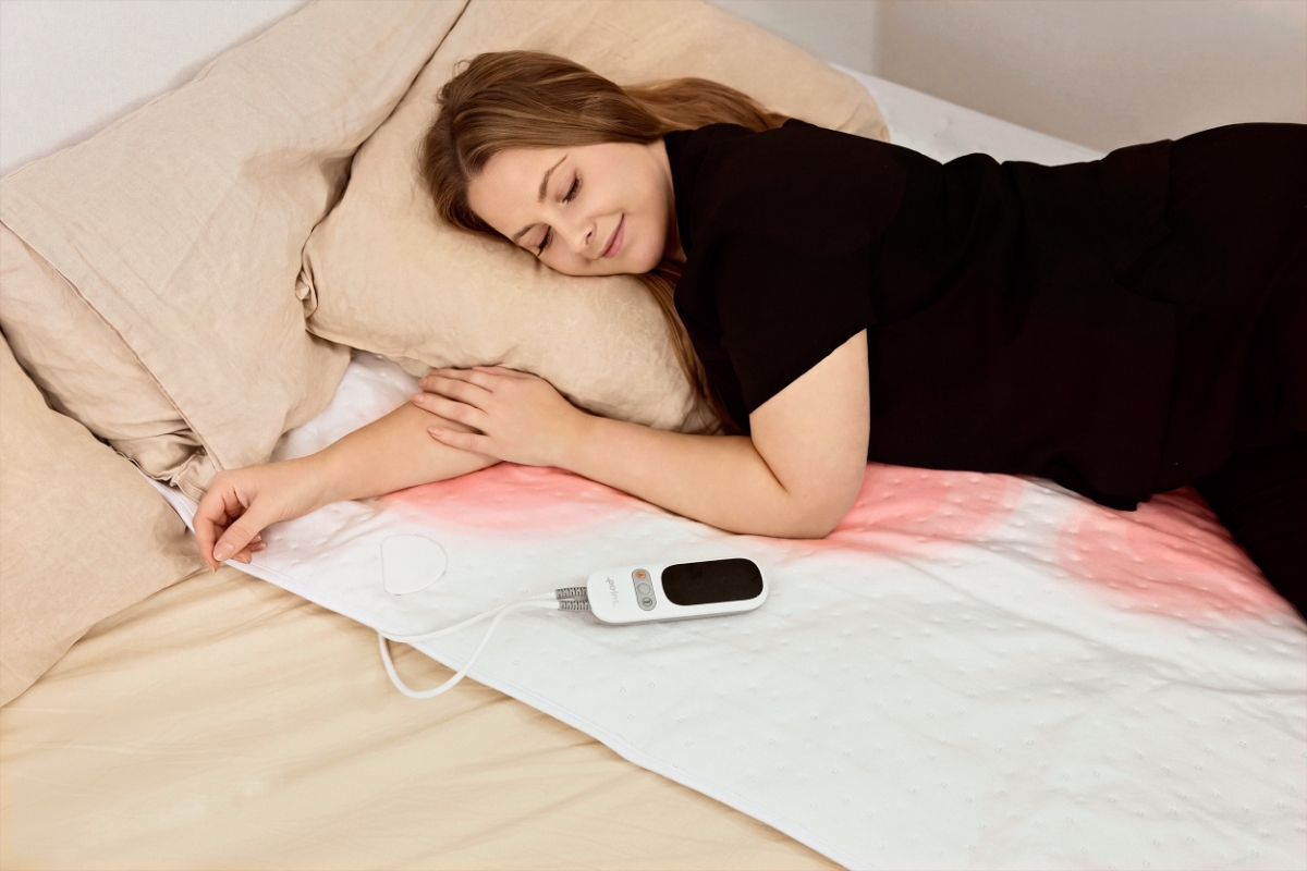 A white single-usable Point heated underblanket on a bed with a woman in a black shirt lying on the bed with red dots under her showing the heated areas on the blanket