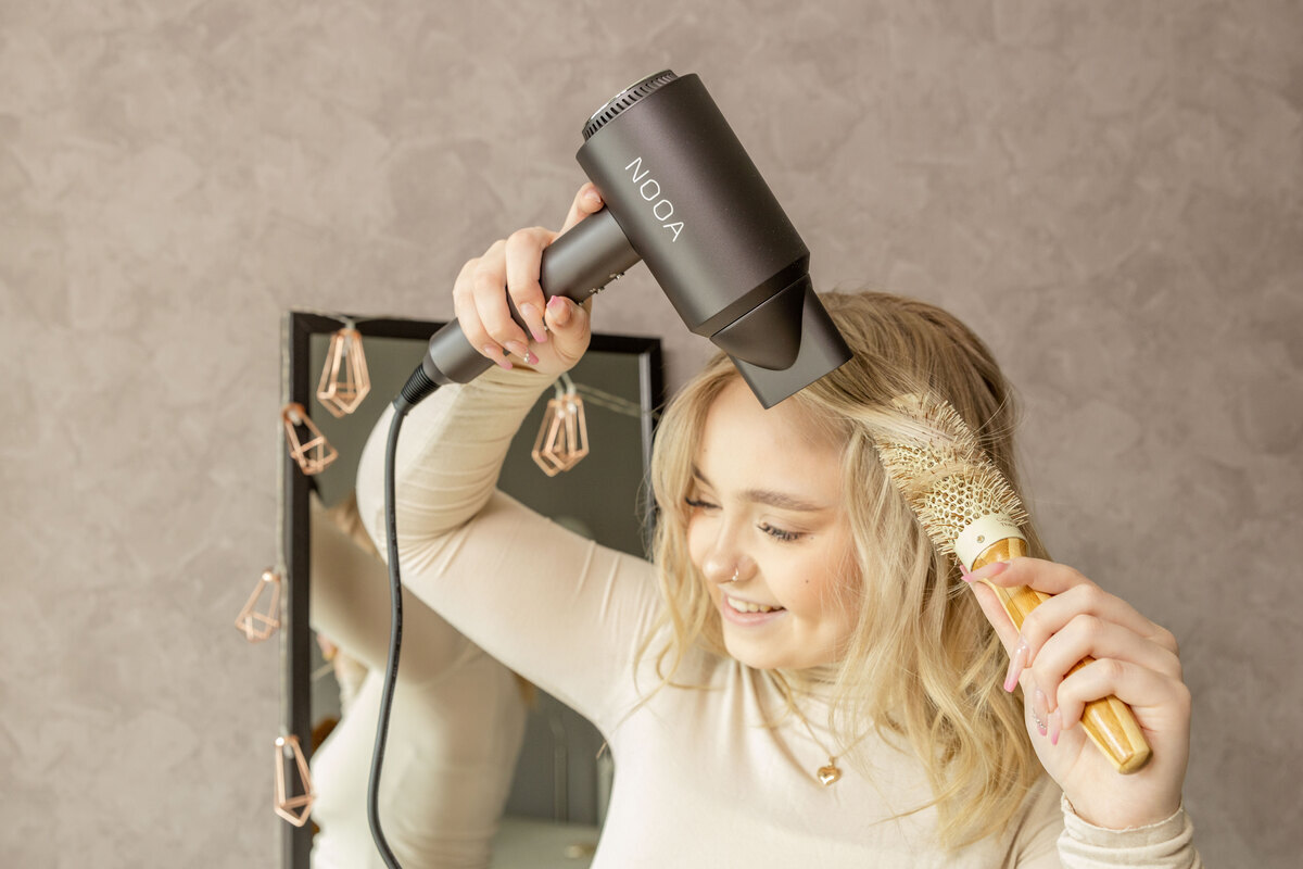 A person wearing a white top and with blonde long hair is curling it using a round brush and the NOOA LUXE BLDC hair dryer in front of a mirror