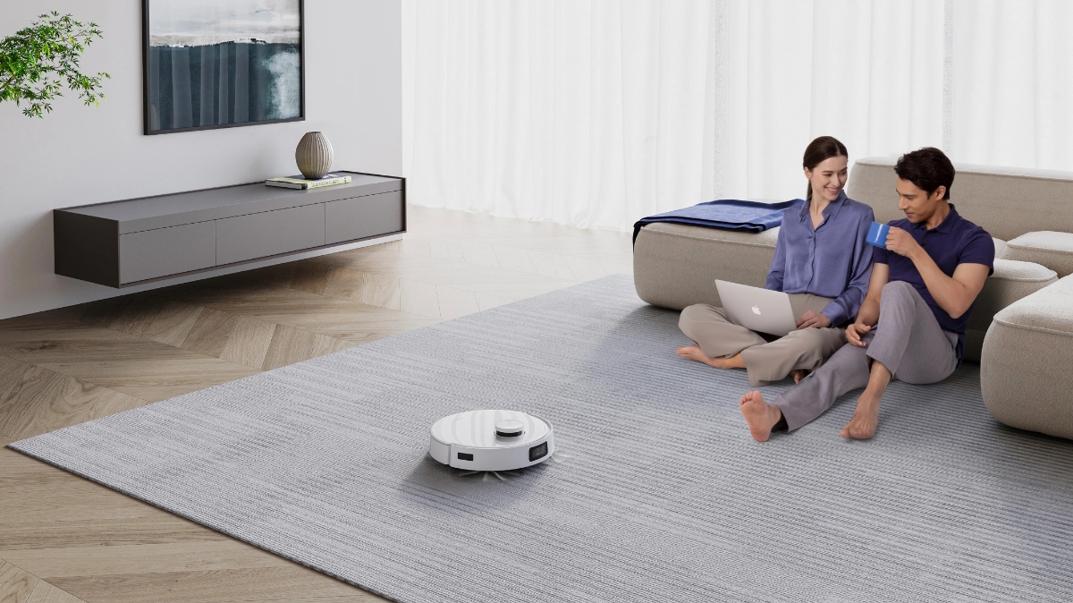 An Ecovacs robotic vacuum cleaner cleaning a big carpet in the living room with two people sitting next to it on the carpet