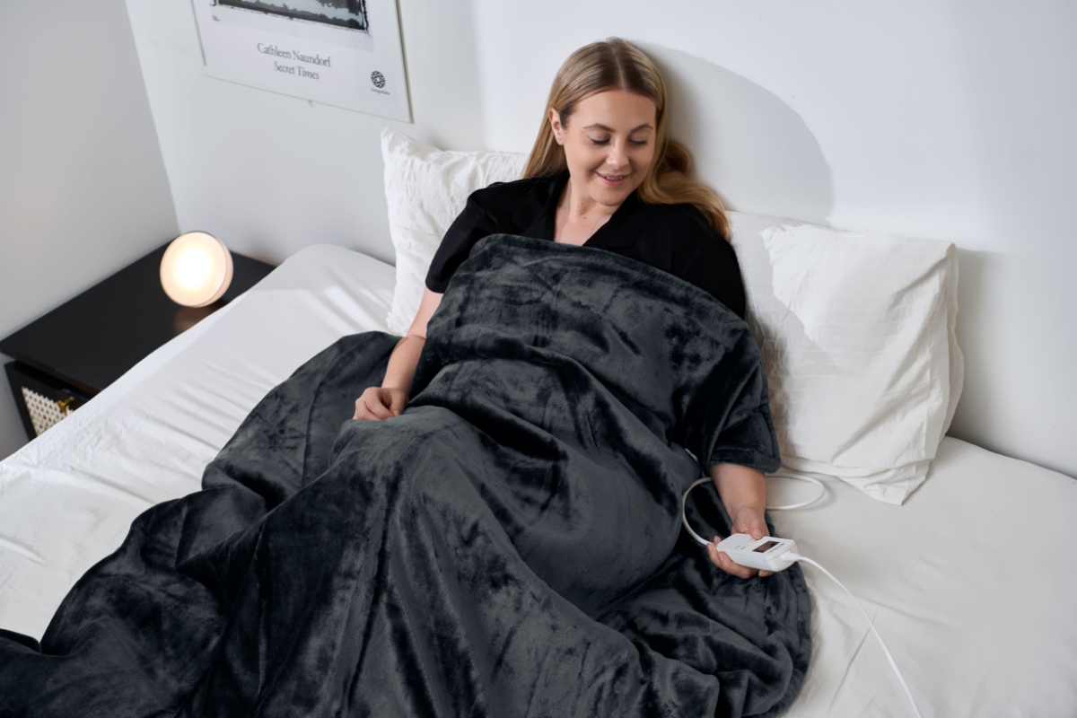 Wide angle image of a person laying under the POINT SOOTHING HEAT HEATING BLANKET, adjusting its setting from the remote control