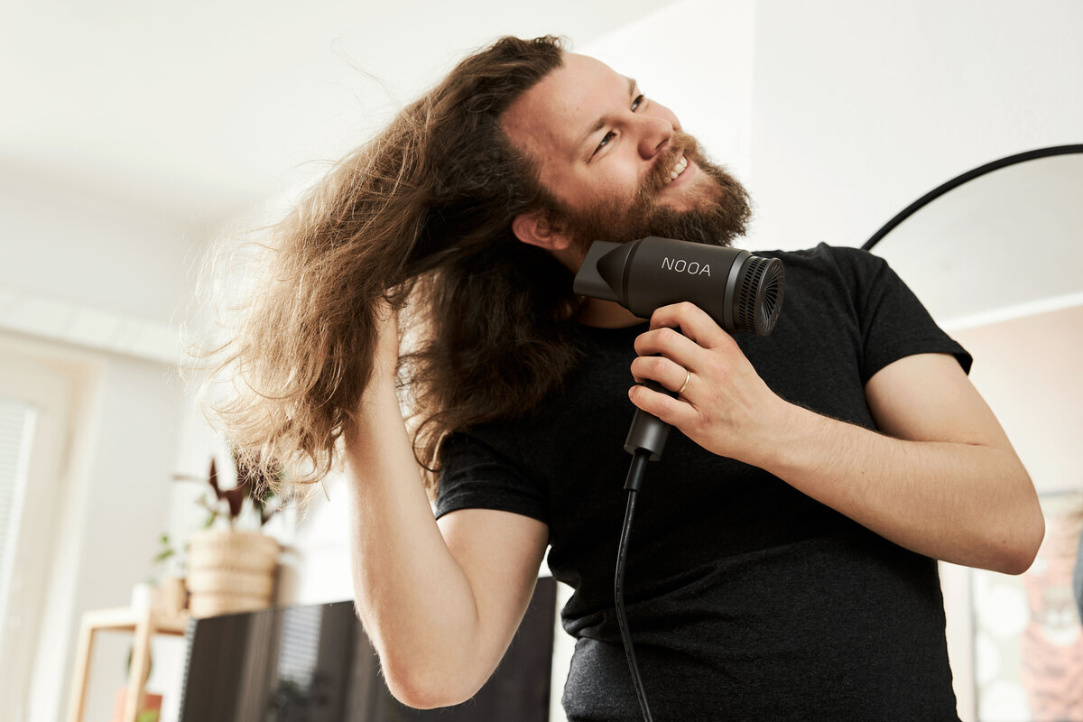 A person wearing a black t shirt and with long thick brown hair is drying it with the NOOA LUXE BLDC hair dryer 