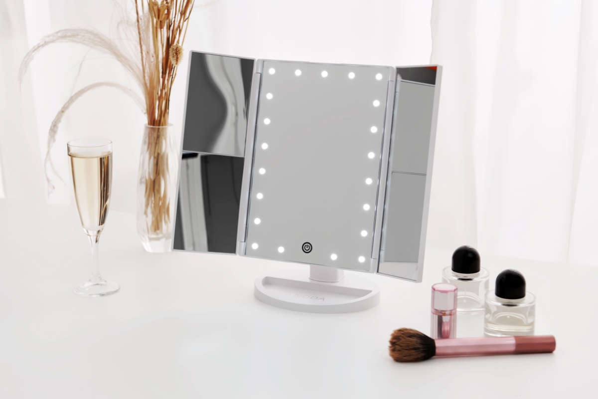 A Nooa Glare make-up mirror standing on a white table with both its side mirror opened and its light on and perfume bottles and a glass of champagne next to the mirror on the table