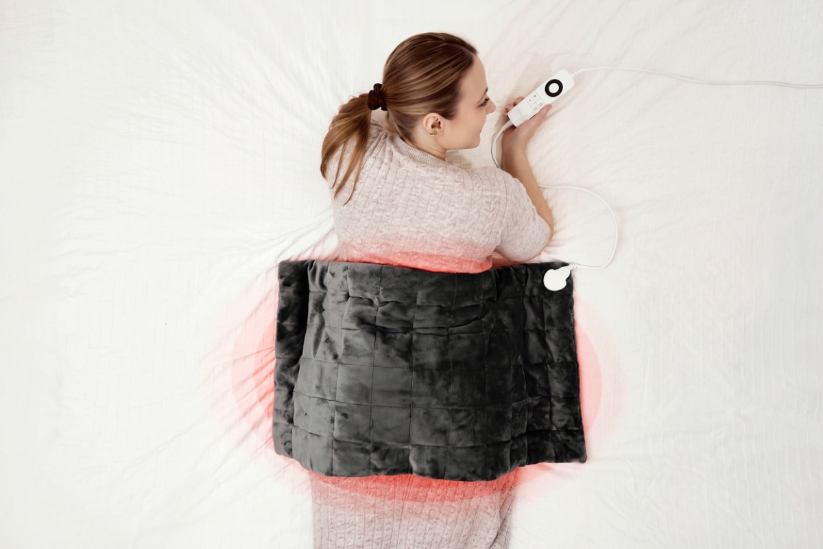 A dark grey Point Lune weighted heating blanket on top of the lower back of a woman lying on a white bed in a beige knitted dress and holding the controller in her hand and looking at the right side smiling