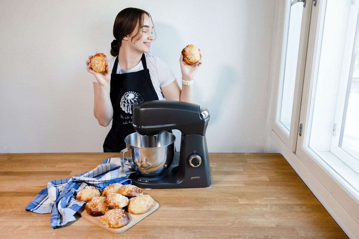 A picture of a grey kitchen machine on a wooden table next to fresh buns and a woman holding the buns in both hands