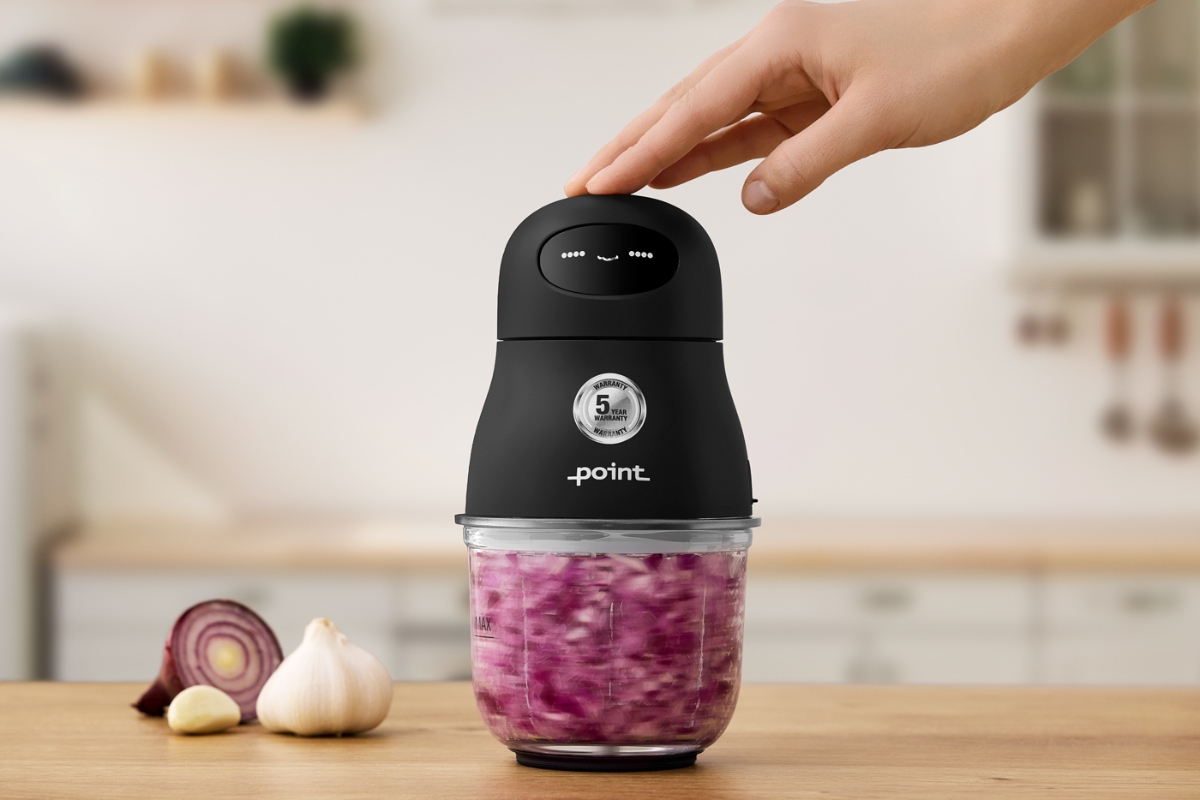 A picture of a black Point cordless food chopper on a wooden table filled with chopped red onions with a light-colored kitchen in the background and a finger pressing the lid of the device