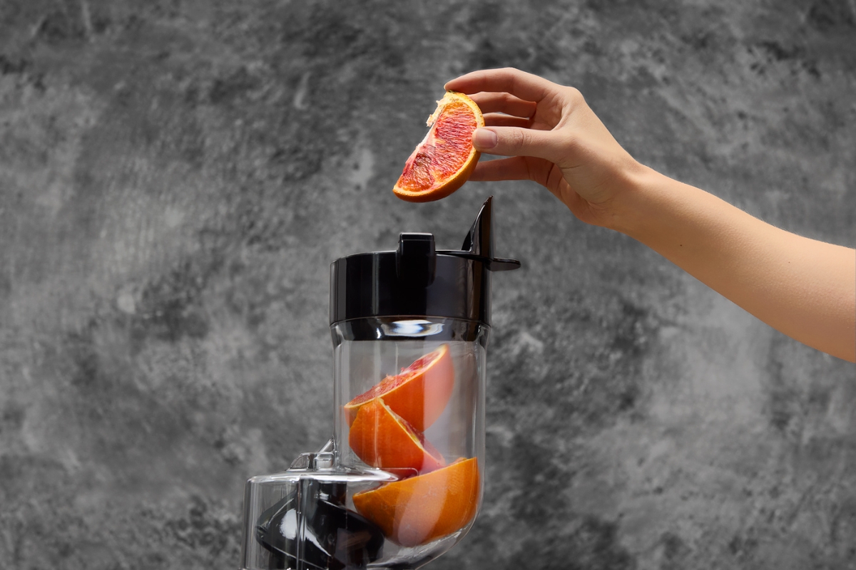 A grey-colored Point slowjuicer against a grey marble wall with its feeding tube pictured up close and a hand putting slices of orange into the tube