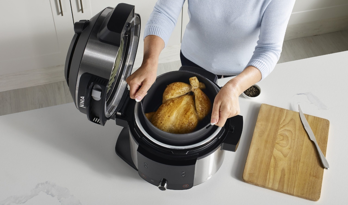 A picture of a woman lifting a roasted chicken out of a black and silver Ninja multicooker that is standing on a white kitchen table