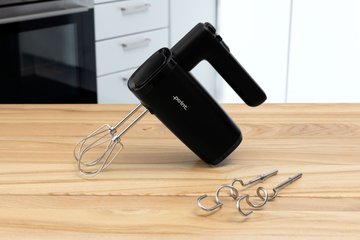 A black Point hand mixer on a wooden table with white appliances behind it and whisks attached to its body and dough hooks next to it on the table