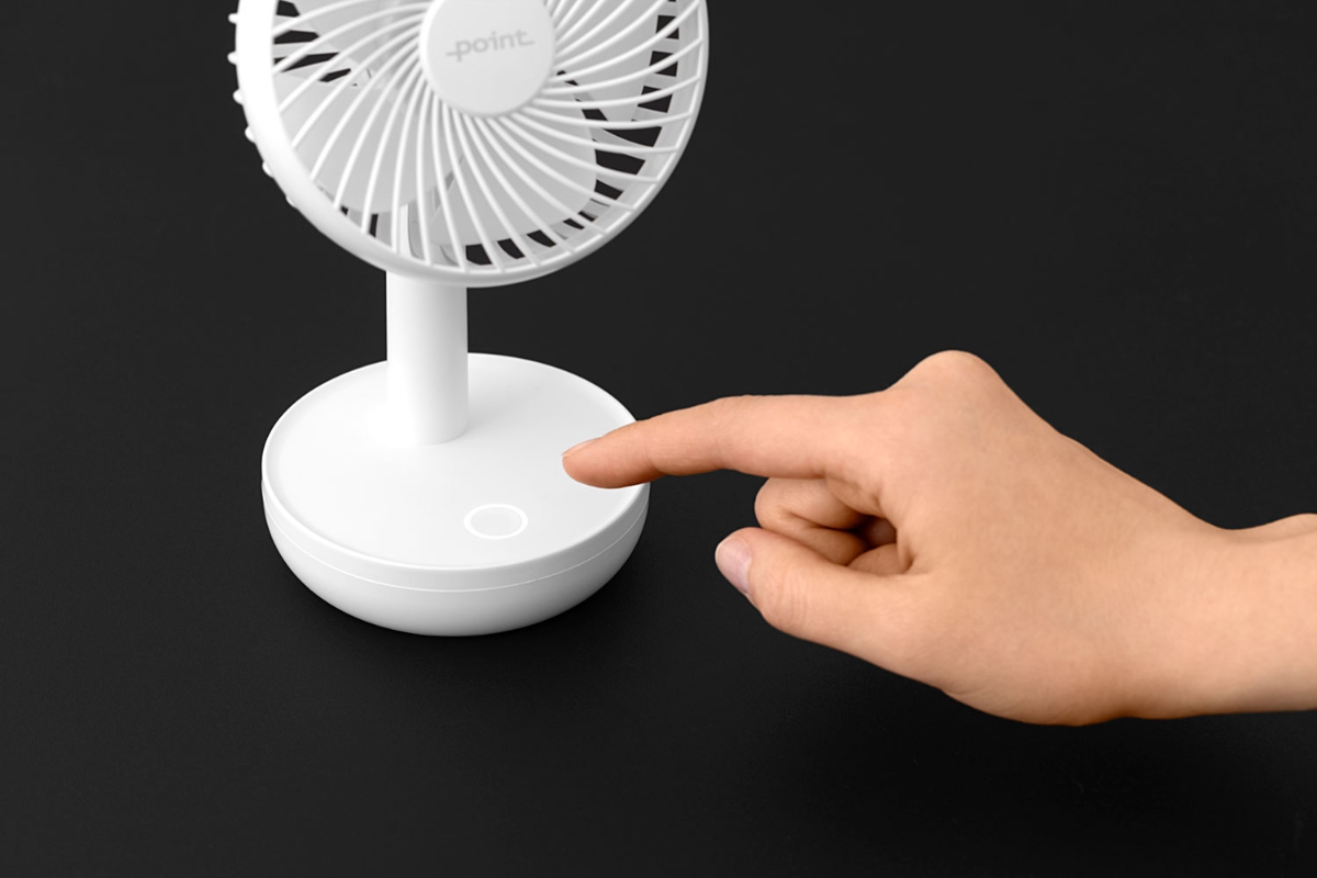 A white and small Point portable table fan with a completely black background and a person pressing the control know on the pedestal with one finger