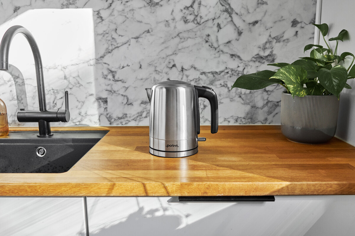 Wide angle image of POINT POKE50SS1 kettle on a wooden kitchen worktop with a plant on the right and a marble wall behind it