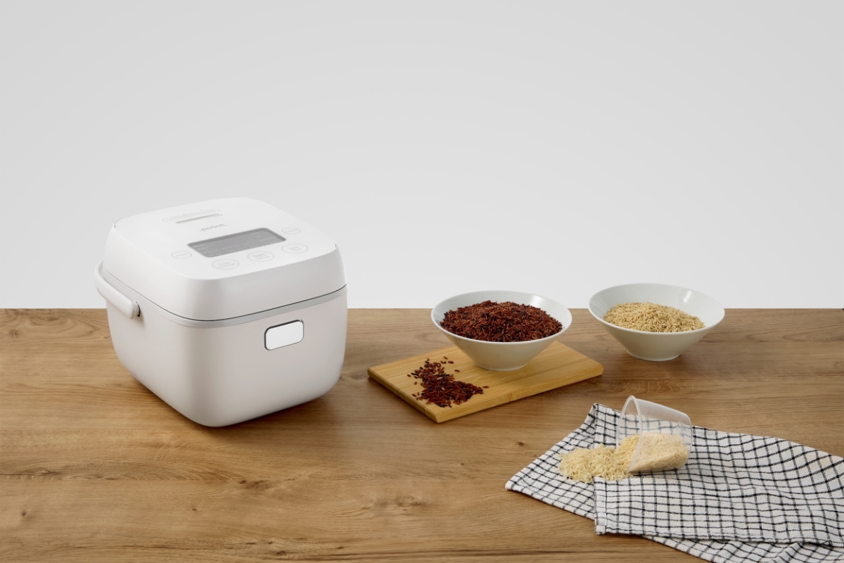 A white Point rice cooker on a wooden table with the machine slightly tilted to the right and different kind of rices next to the machine on the table on wooden chopboards and kitchen linens