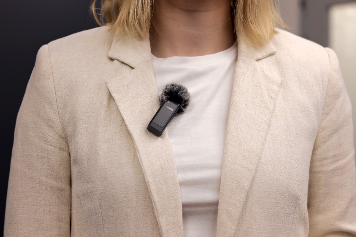 En sort tr&aring;dl&oslash;s mikrofon med lodden vindhette festet p&aring; jakkeslaget til en kvinne if&oslash;rt en kremfarget blazer over en hvit t-skjorte.