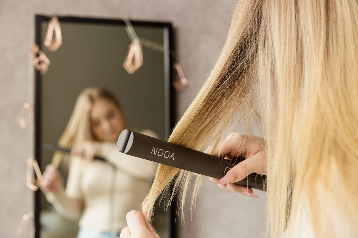 A woman straightening her blonde hair in front of a mirror with a grey NOOA hair straightener