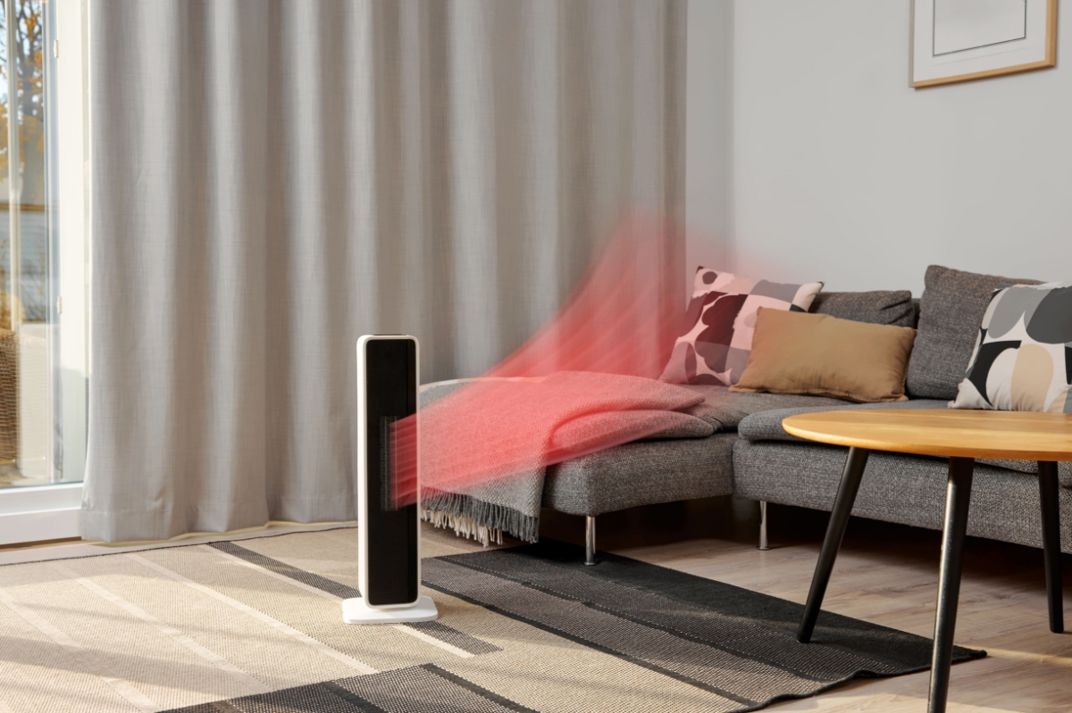 A black Point fan heater standing in a living room with beige and black carpet and a grey sofa next to the heater and light-grey curtains behind the appliance and red heat coming out of the device