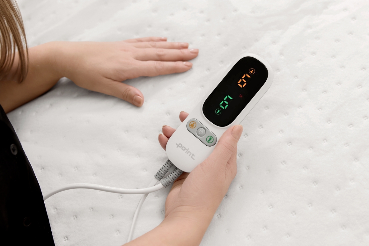 A white Point heated underblanket on the bed and a woman with neat nails and a black shirt holding the controller of the blanket in her hand and the other hand touching the blanket
