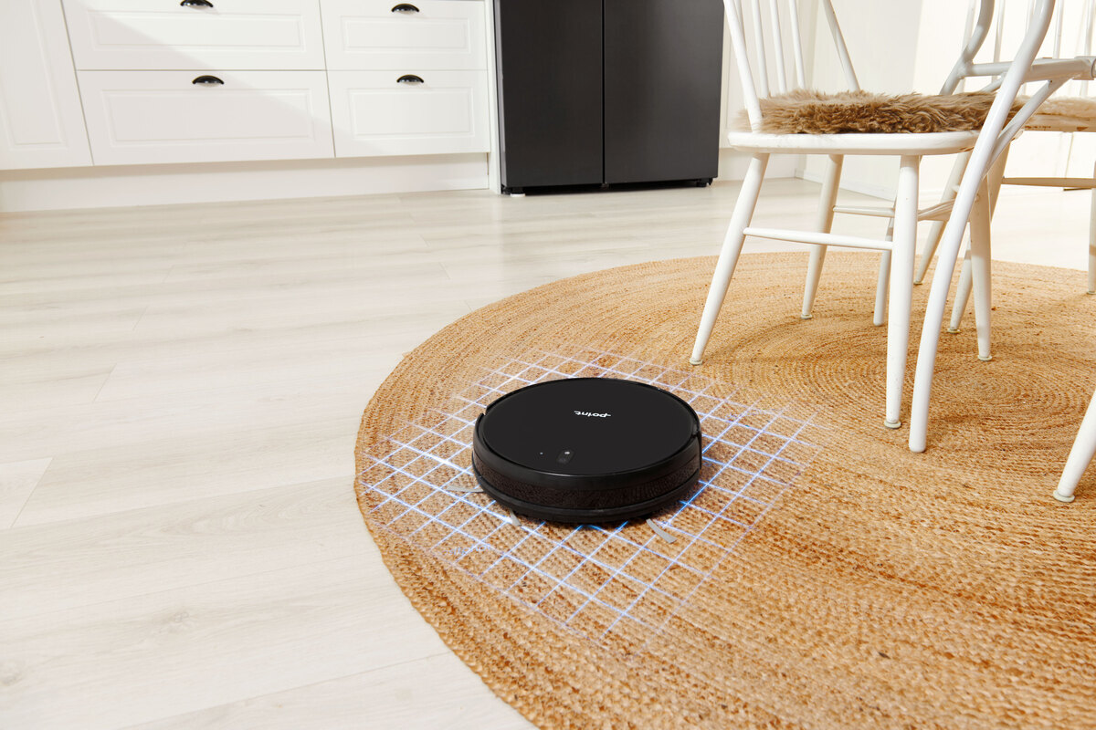 A black Point robotic vacuum cleaner vacuuming a light brown carpet next to white kitchen chairs and white kitchen cabinets on the background