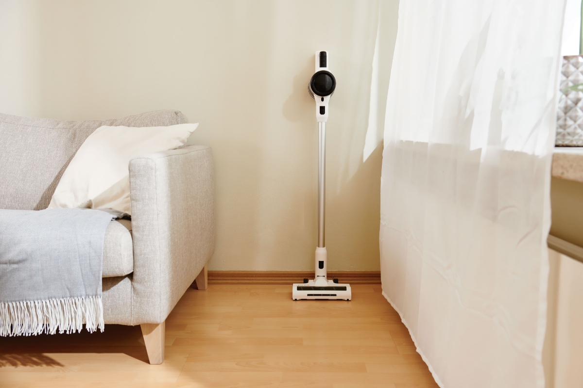 A wide angle image of the Point Pencil stick vacuum in front of a beige wall with a white sofa next to it