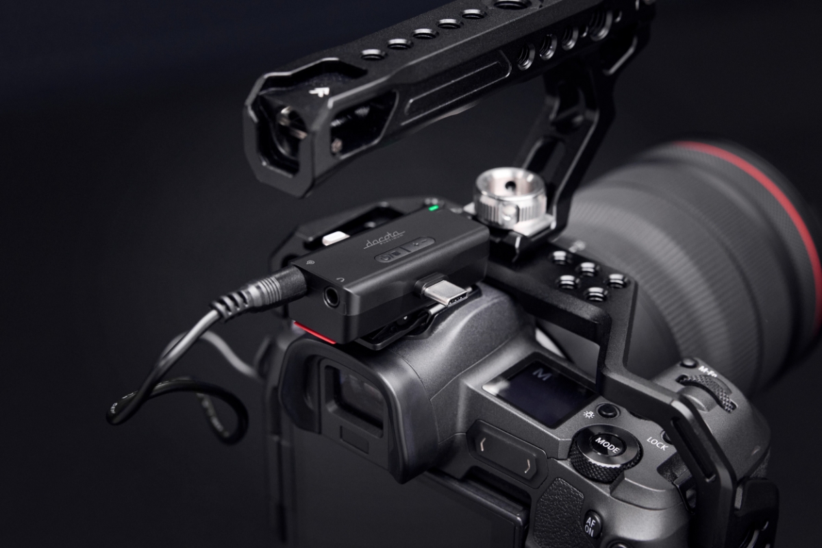 N&aelig;rbilde av en svart Dacota mikrofonadapter koblet til et digitalkamera med en svart kabel. Kameraet er utstyrt med et topph&aring;ndtak og en linse synlig i bakgrunnen.