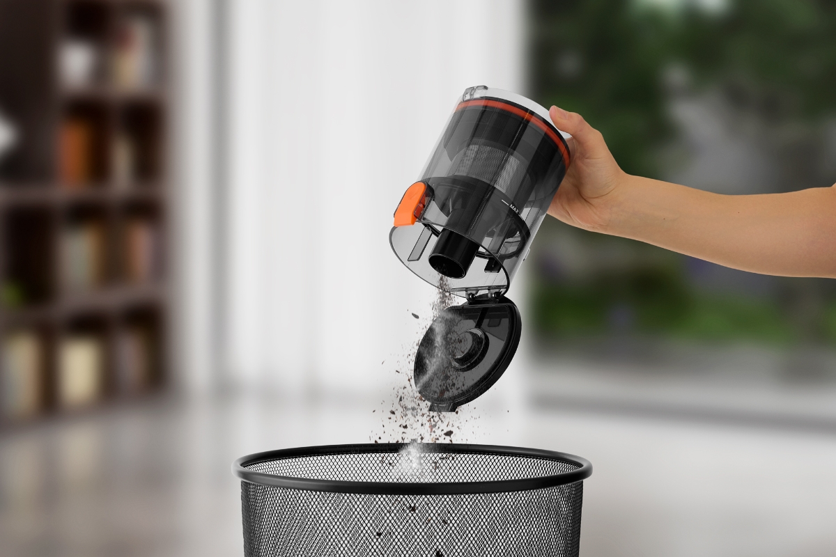 A black and red colored Point Cruise stick vacuum cleaner's dust container being emptied to a trash can with dust pouring out of the container and a bright room in the background