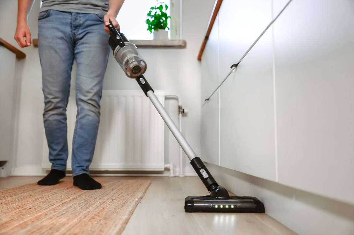 Close up of the POINT POVC618DB 18V stick vacuum vacuumin underneath light grey kitchen cabinets with the LED lights on it, person with blue jeans operating the vacuum