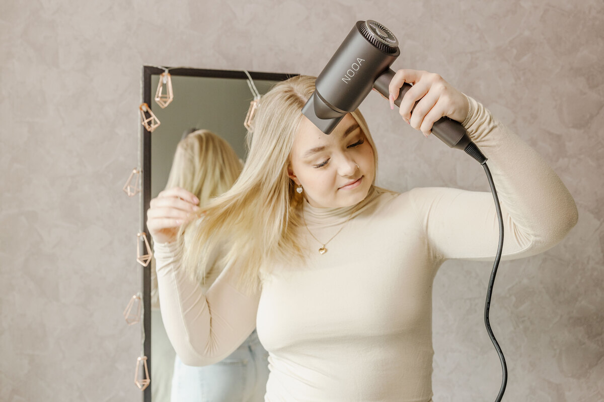 A wide image of a blonde person wearing a white top drying their hair with the NOOA LUXE BLDC hair dryer in front of a mirror