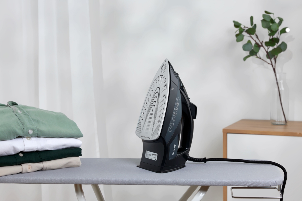 A picture of a black Point steam iron in an upright position on an ironing board with a plant behind the device and a pile of clothes on the board