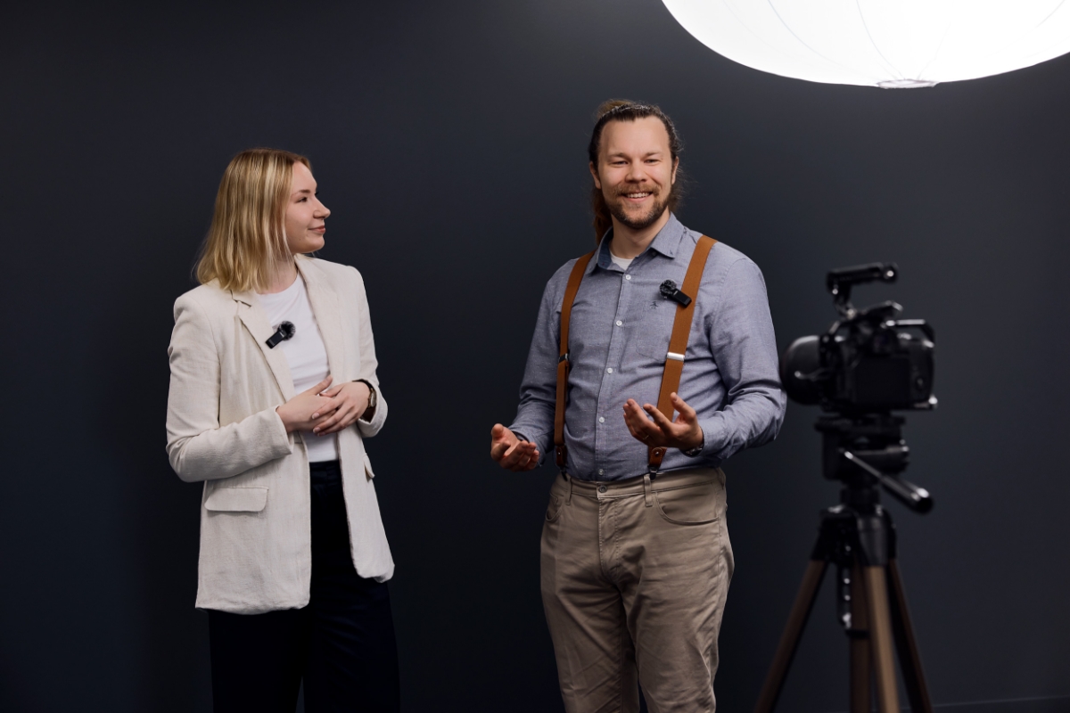 Livsstilsbilde med to personer som bruker tr&aring;dl&oslash;se clip-on mikrofoner, plassert foran et kamera p&aring; et stativ, mot en m&oslash;rk bakgrunn med myk belysning ovenfra.