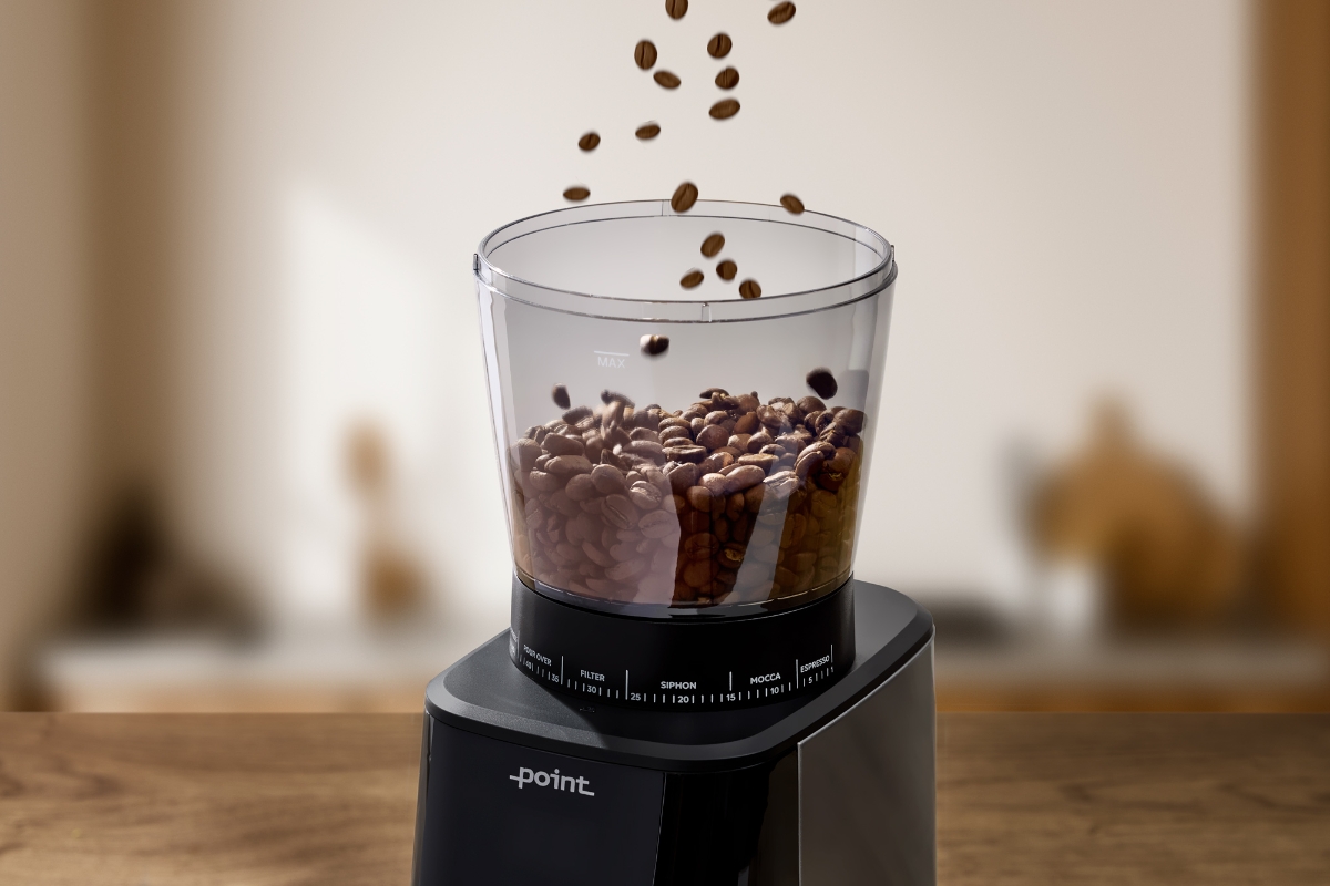 A black Point Pro coffee grinder pictured up close with its coffee bean container being filled with dropping coffee beans and a wooden kitchen showing in the background blurry