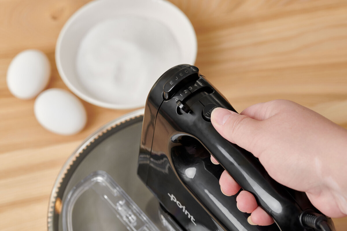 A close-up picture of a black and steel hand mixer with a person pressing its settings and two eggs on the table next to the appliance